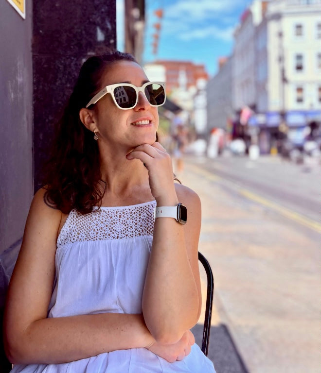 Woman in sunglasses and white dress sitting outdoors, man in light blue shirt and sunglasses standing outdoors.
