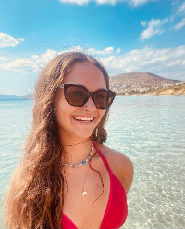 Woman in a red bikini and sunglasses at a beach with clear water and blue sky.